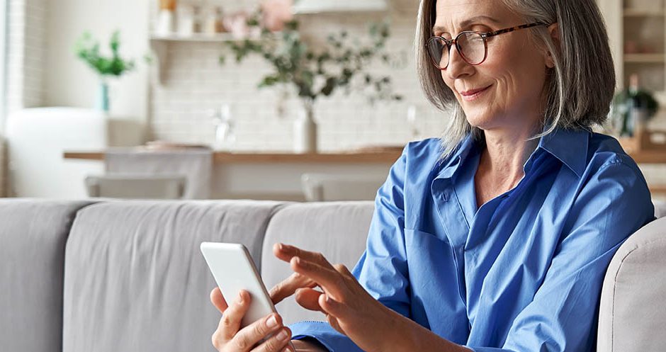Relaxed mature old 60s woman, older middle aged female customer holding smartphone using mobile app, texting message, search ecommerce offers on cell phone technology device sitting on couch at home.